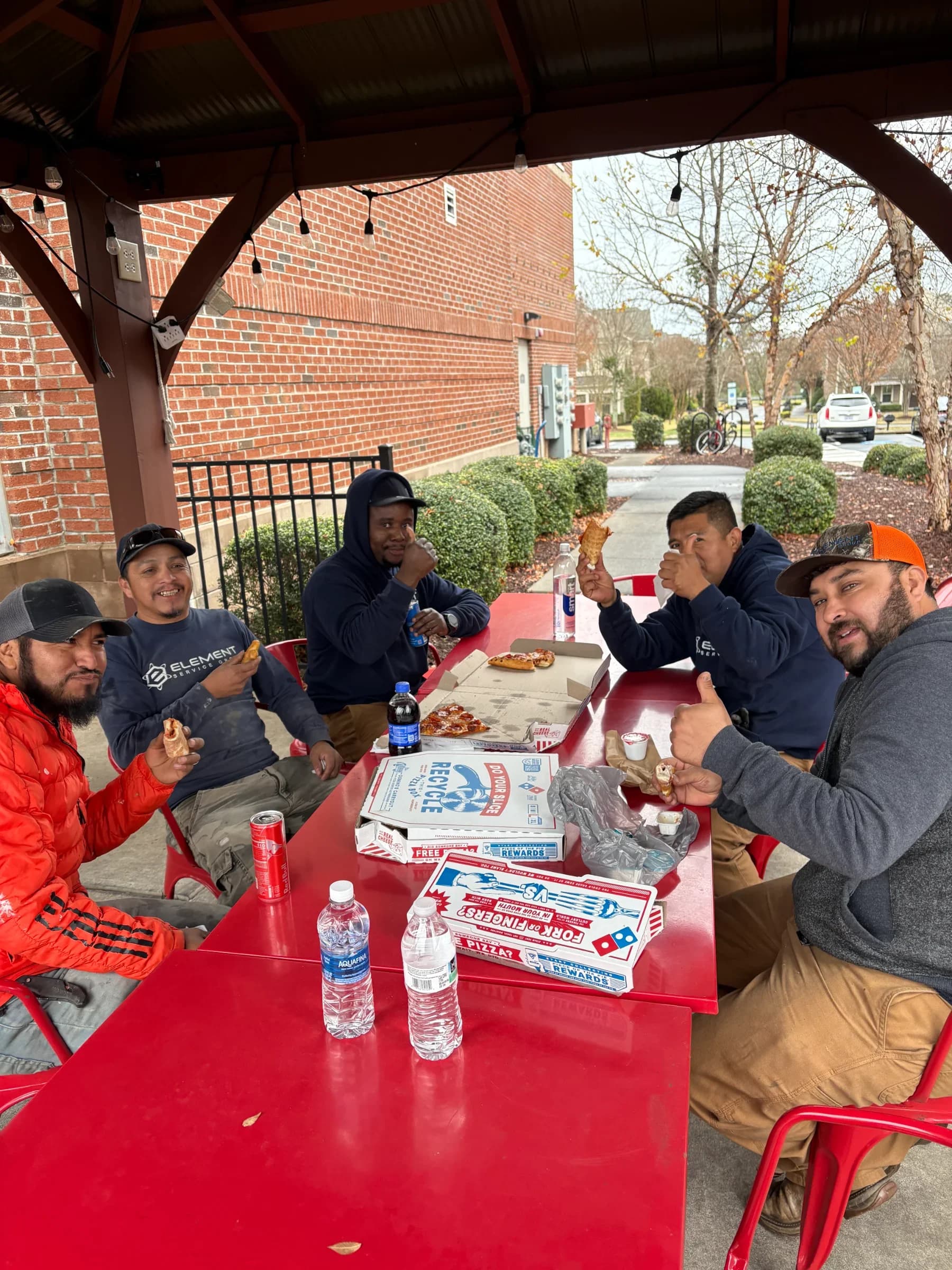 HVAC plumbing team enjoying pizza lunch outdoors at a red table in a work environment